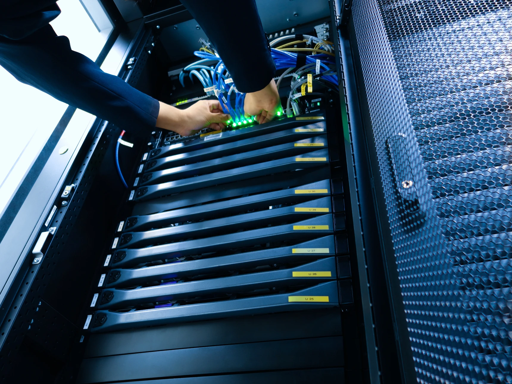 Photographie d'un informaticien professionnel en tenue de travail effectuant une intervention de maintenance informatique : il ajuste ou inspecte du matériel serveur dans une baie informatique, concentré sur sa tâche, dans un environnement de datacentre ou de salle serveur bien éclairé et organisé. L'image doit transmettre la confiance, la compétence technique et le professionnalisme, illustrant le maintien en condition opérationnelle de l'infrastructure informatique d'une entreprise cliente.